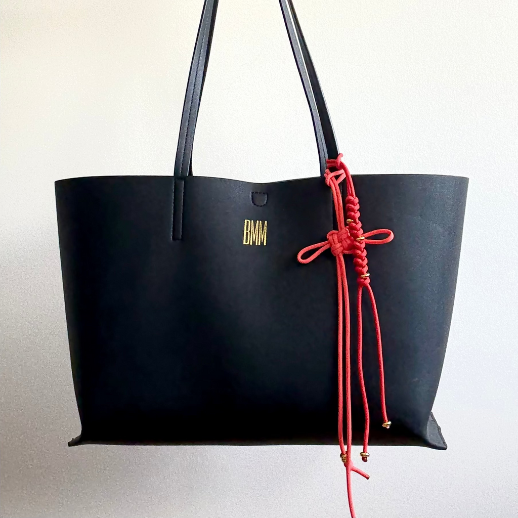 Black tote bag with red cord and gold logo on a light gray background